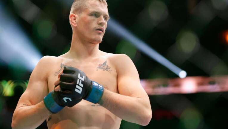 Male mixed martial artist in blue UFC gloves stands in a fighting stance inside the cage, focused and shirtless, with tattoos on his chest.