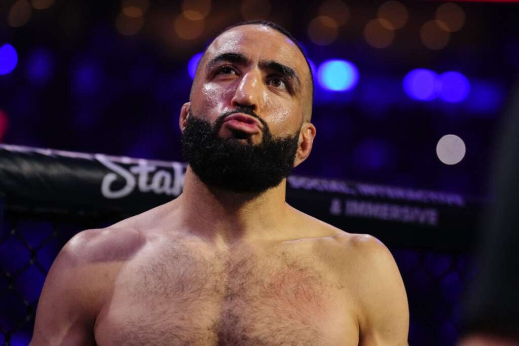 Bearded, shirtless MMA fighter in the octagon, looking up with a determined expression as blue arena lights glow behind him.