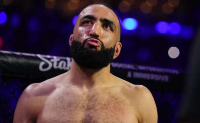 Bearded, shirtless MMA fighter in the octagon, looking up with a determined expression as blue arena lights glow behind him.