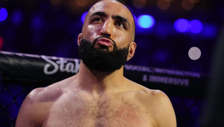 Bearded, shirtless MMA fighter in the octagon, looking up with a determined expression as blue arena lights glow behind him.