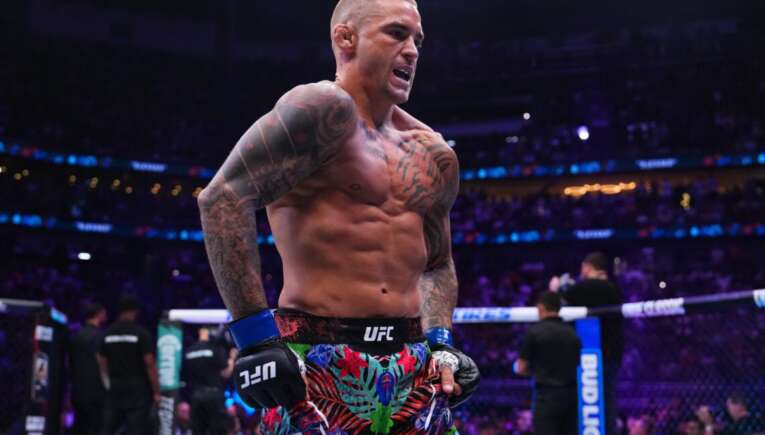 UFC fighter with tattoos and colorful floral shorts standing in the octagon, shirtless and looking intense as the crowd watches.