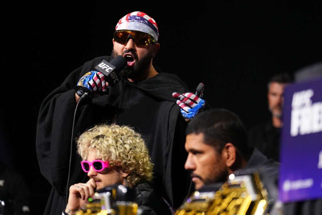 UFC fighter wearing a patriotic bandana and sunglasses speaks into a microphone on stage, gesturing with gloved hands.