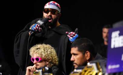 UFC fighter wearing a patriotic bandana and sunglasses speaks into a microphone on stage, gesturing with gloved hands.