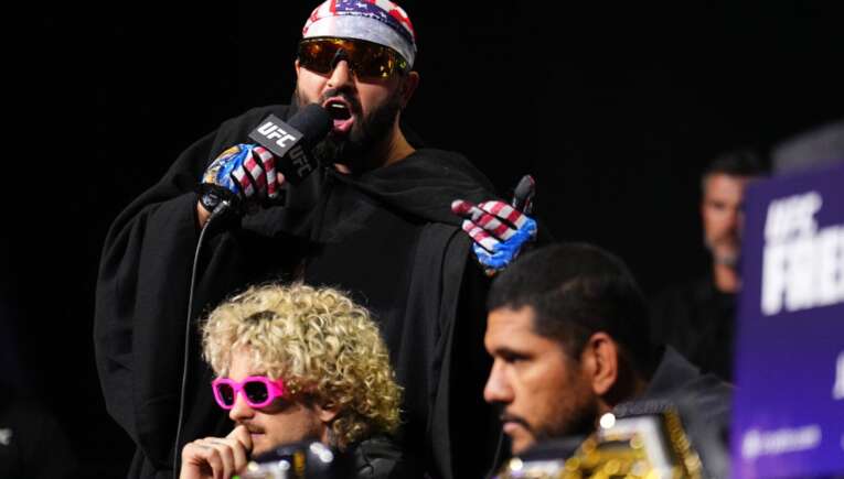 UFC fighter wearing a patriotic bandana and sunglasses speaks into a microphone on stage, gesturing with gloved hands.