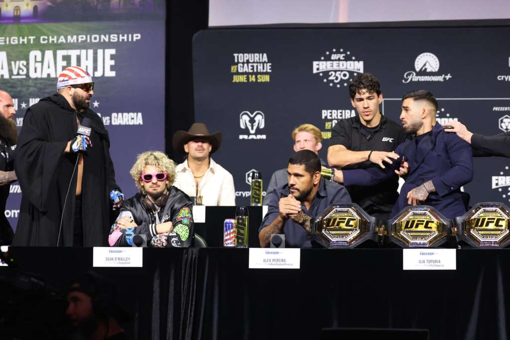 UFC press conference: a man in a black cloak speaks into a microphone at a table with championship belts in front, backdrop branding behind him.