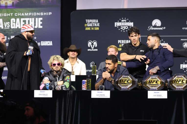 Group of UFC fighters seated behind a long table with championship belts, reporters and banners in the background at a press conference.