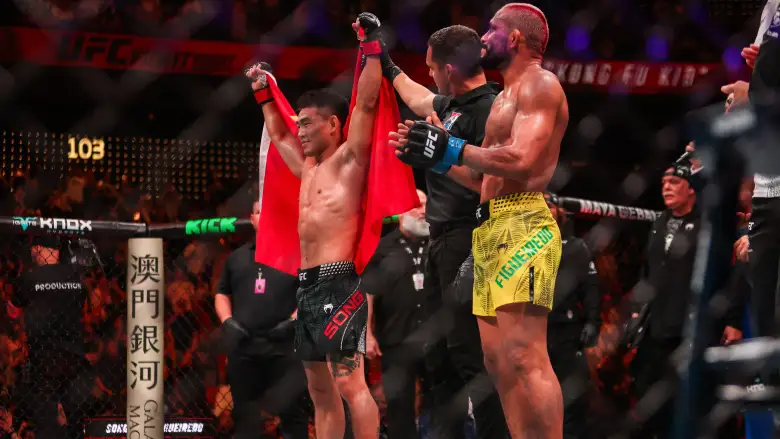 Two UFC fighters celebrate inside the cage as a referee raises both arms after a win, one in red shorts and cape and the other in yellow shorts, with a cheering crowd in the background
