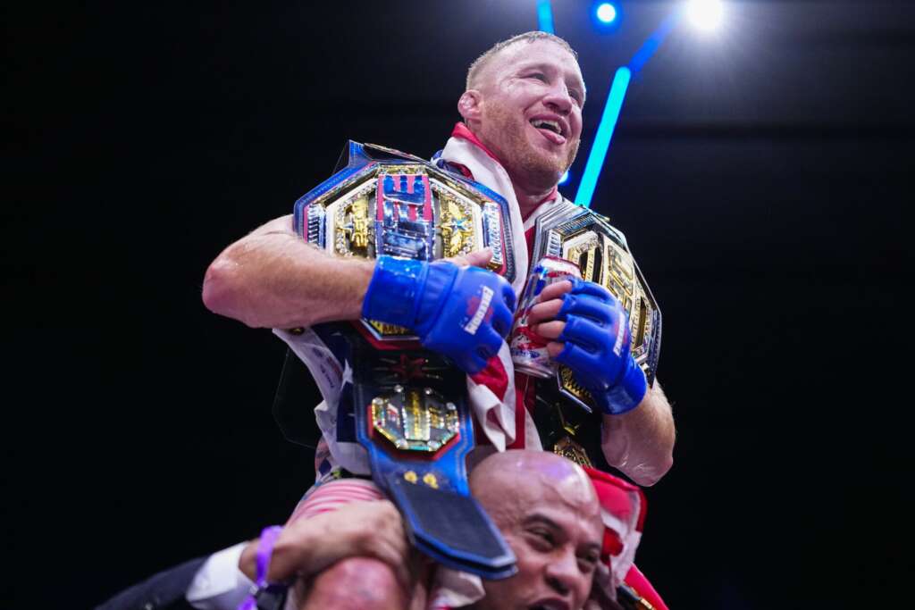 A victorious boxer wearing multiple championship belts and blue gloves celebrates while being lifted by a teammate under bright arena lights.