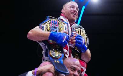 A victorious boxer wearing multiple championship belts and blue gloves celebrates while being lifted by a teammate under bright arena lights.