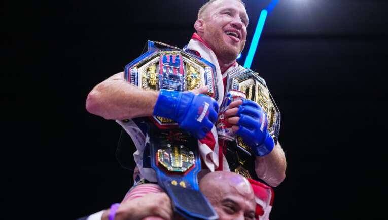 A victorious boxer wearing multiple championship belts and blue gloves celebrates while being lifted by a teammate under bright arena lights.