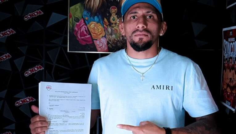 Man standing indoors, holding a printed document in his left hand and pointing to it with his right hand, wearing a light blue shirt and cap, against a wall with art and posters behind him.