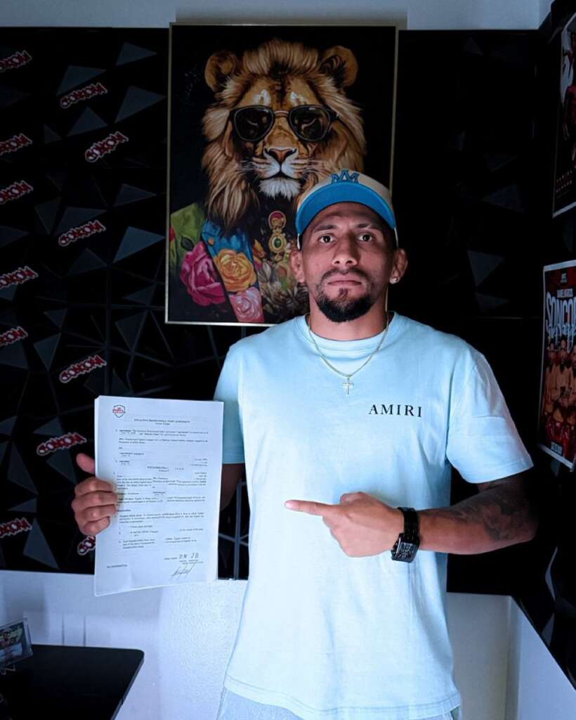 Man standing indoors, holding a printed document in his left hand and pointing to it with his right hand, wearing a light blue shirt and cap, against a wall with art and posters behind him.