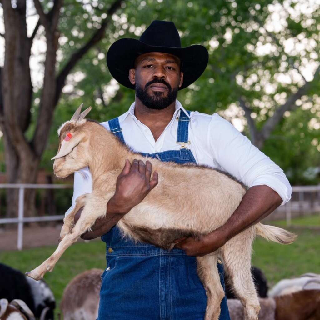 Man wearing a black cowboy hat and denim overalls holds a light-colored goat kid in an outdoor farm setting with trees and fencing behind him.