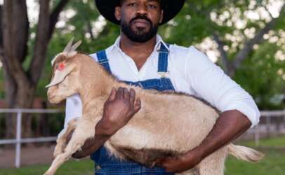 Man wearing a black cowboy hat and denim overalls holds a light-colored goat kid in an outdoor farm setting with trees and fencing behind him.