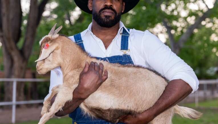 Man wearing a black cowboy hat and denim overalls holds a light-colored goat kid in an outdoor farm setting with trees and fencing behind him.