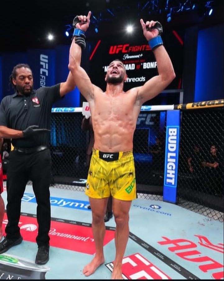 Oplus_131072 - AfroCombat Mixed martial arts fighter in yellow shorts raises both arms in victory inside the UFC octagon, with the referee standing beside him and crowd lights in the background.