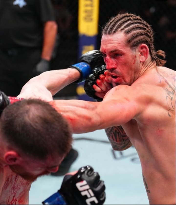 Two MMA fighters clash in the octagon as one lands a punch on the other, both bloodied with taped hands and tattoos visible on their bodies.