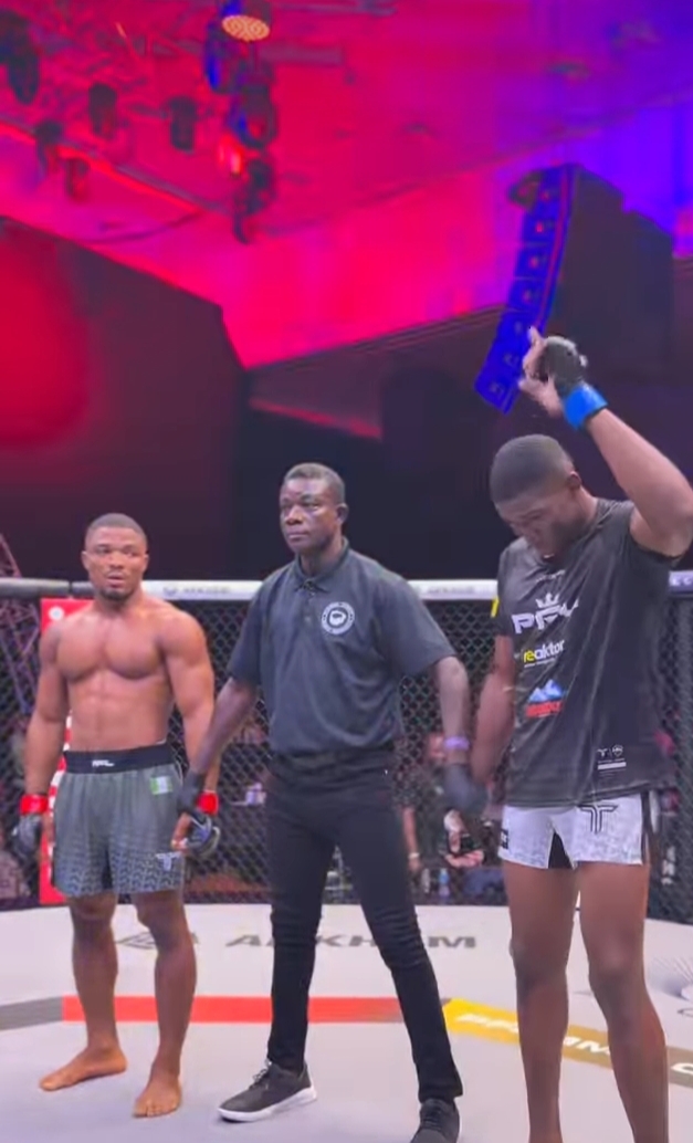 Post-fight moment in a cage: referee between two shirtless fighters; the right fighter raises a blue championship belt in victory.
