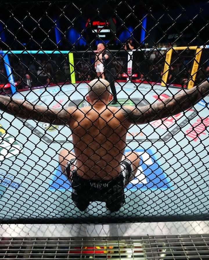 Back view of a shirtless fighter seated with arms resting on the chain-link fence inside a mixed martial arts cage before a bout.