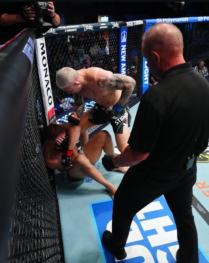 MMA fight inside a cage: one fighter on the mat defending while the standing opponent leans over, a referee watches from the foreground.