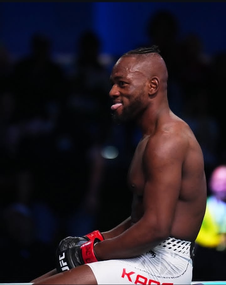 Male mixed martial artist sitting in the ring, shirtless, wearing white shorts and red gloves, smiling with tongue out.