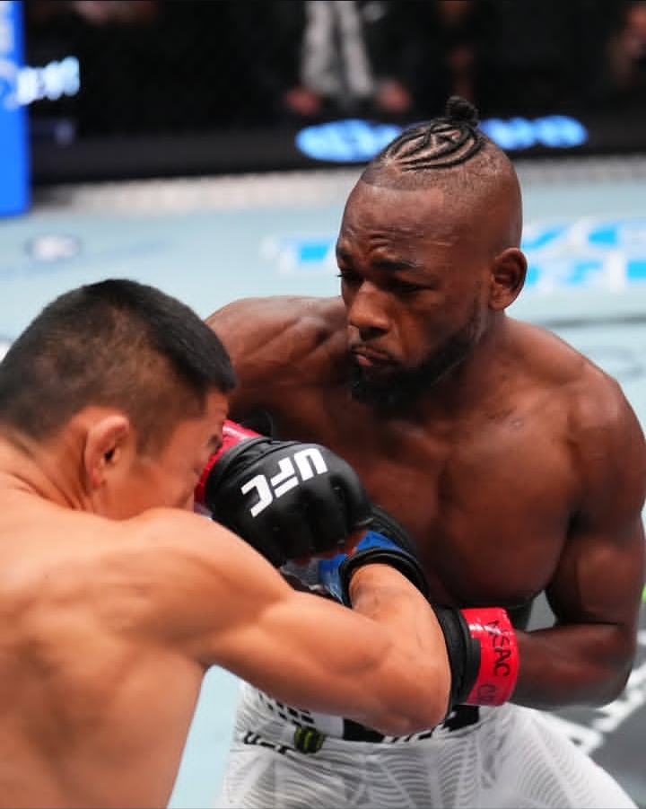 Two UFC fighters clash in the cage; one lands a punch while the other braces his guard with red gloves and blue shorts visible.