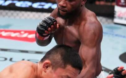 MMA fight moment: a muscular Black fighter in white shorts lands a strike on an opponent crouched in the cage.