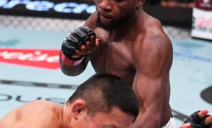 MMA fight moment: a muscular Black fighter in white shorts lands a strike on an opponent crouched in the cage.