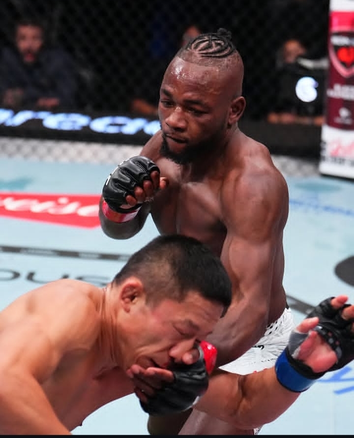MMA fight moment: a muscular Black fighter in white shorts lands a strike on an opponent crouched in the cage.