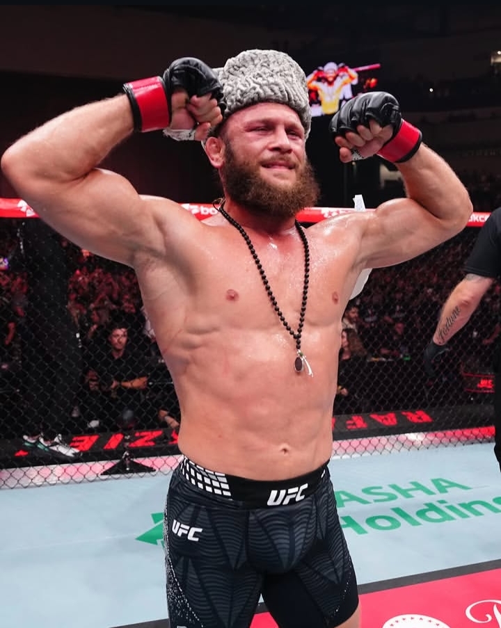 MMA fighter in the octagon flexes arms, smiling after a win, wearing a furry gray beanie and a bead necklace.
