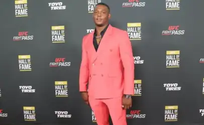 Man wearing a bright pink suit posing on a red carpet with UFC Hall of Fame backdrop behind him