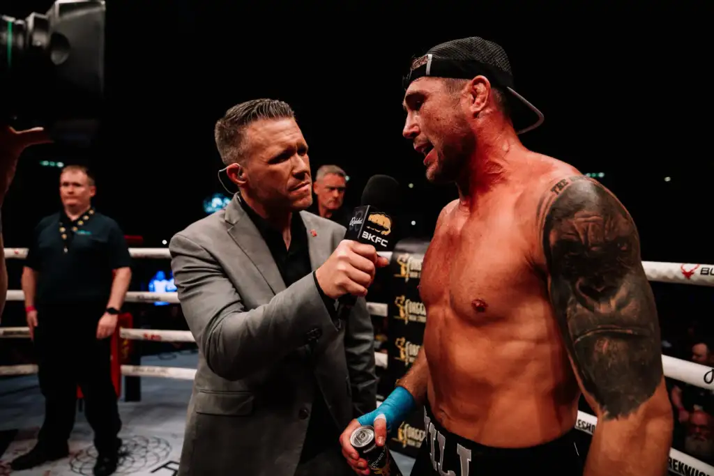Reporter in a gray suit interviews a shirtless, tattooed boxer in the ring, holding a BKFC microphone.