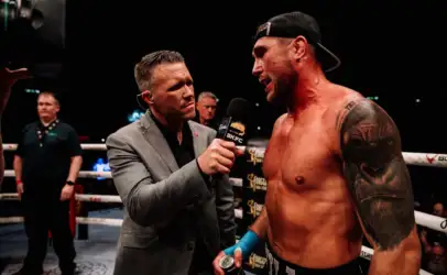 Reporter in a gray suit interviews a shirtless, tattooed boxer in the ring, holding a BKFC microphone.