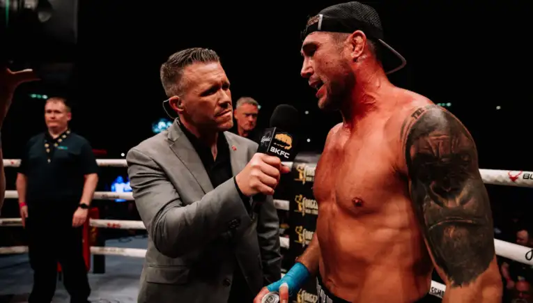 Reporter in a gray suit interviews a shirtless, tattooed boxer in the ring, holding a BKFC microphone.
