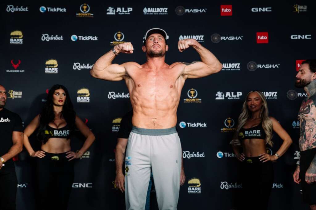 Muscular, shirtless man flexing on a stage in gray shorts, flanked by two promo models in black Bare Knuckle outfits against sponsor logos backdrop.