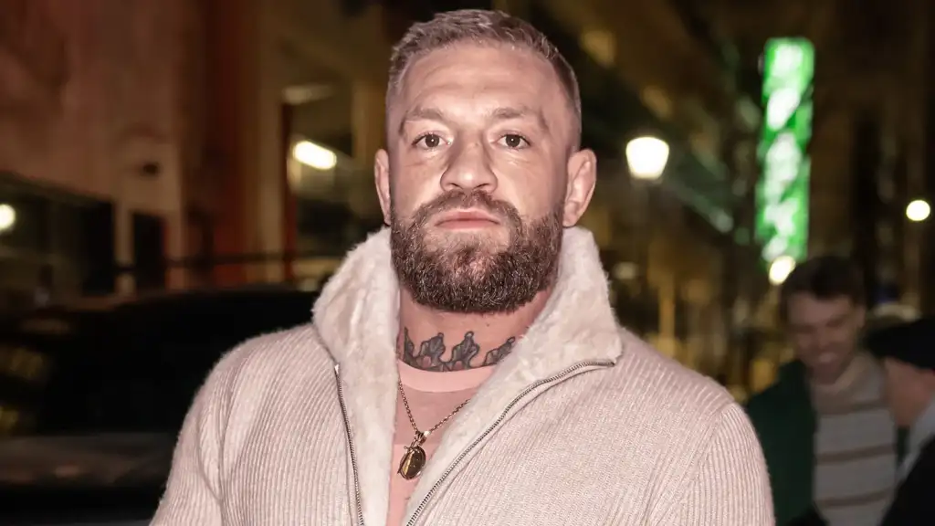 Bearded man with neck tattoo wearing a cream zip-up sweater with a shearling collar, posing on a city street at night.