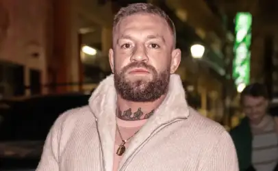 Bearded man with neck tattoo wearing a cream zip-up sweater with a shearling collar, posing on a city street at night.