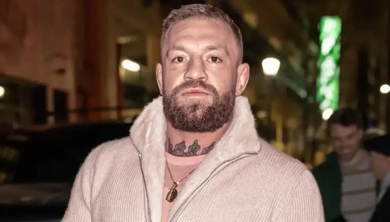 Bearded man with neck tattoo wearing a cream zip-up sweater with a shearling collar, posing on a city street at night.