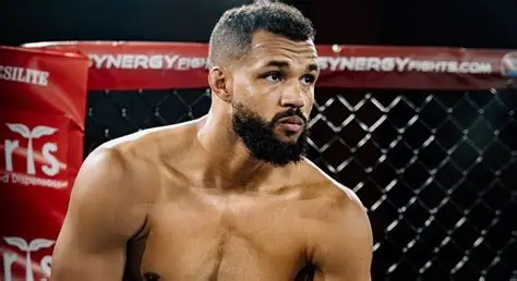 shirtless male MMA fighter in a cage, looking off to the side with a focused expression inside the octagon
