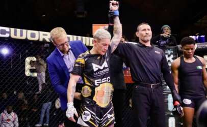 Referee raises the arm of a victorious MMA fighter wearing black-and-gold shorts and a championship belt inside the cage, with a man in a blue suit nearby.