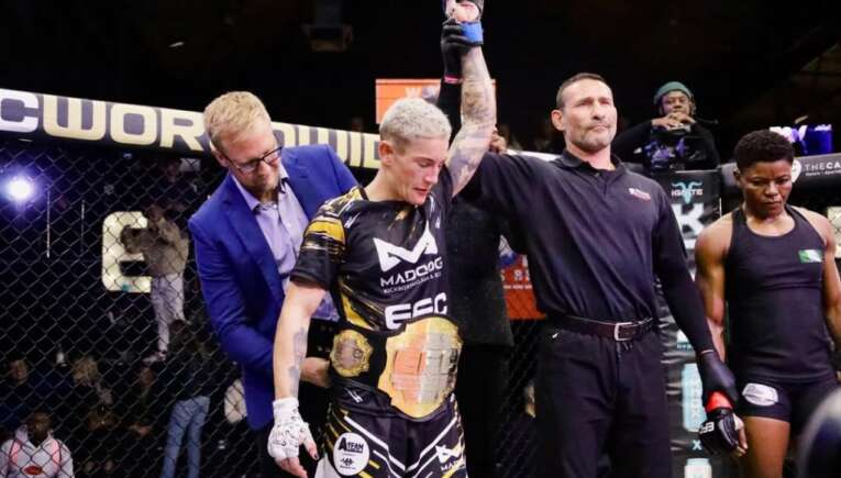 Referee raises the arm of a victorious MMA fighter wearing black-and-gold shorts and a championship belt inside the cage, with a man in a blue suit nearby.