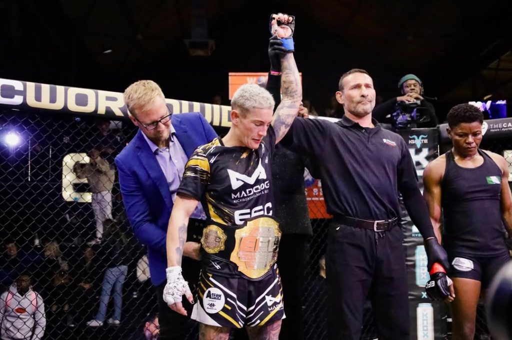 Referee raises the arm of a victorious MMA fighter wearing black-and-gold shorts and a championship belt inside the cage, with a man in a blue suit nearby.