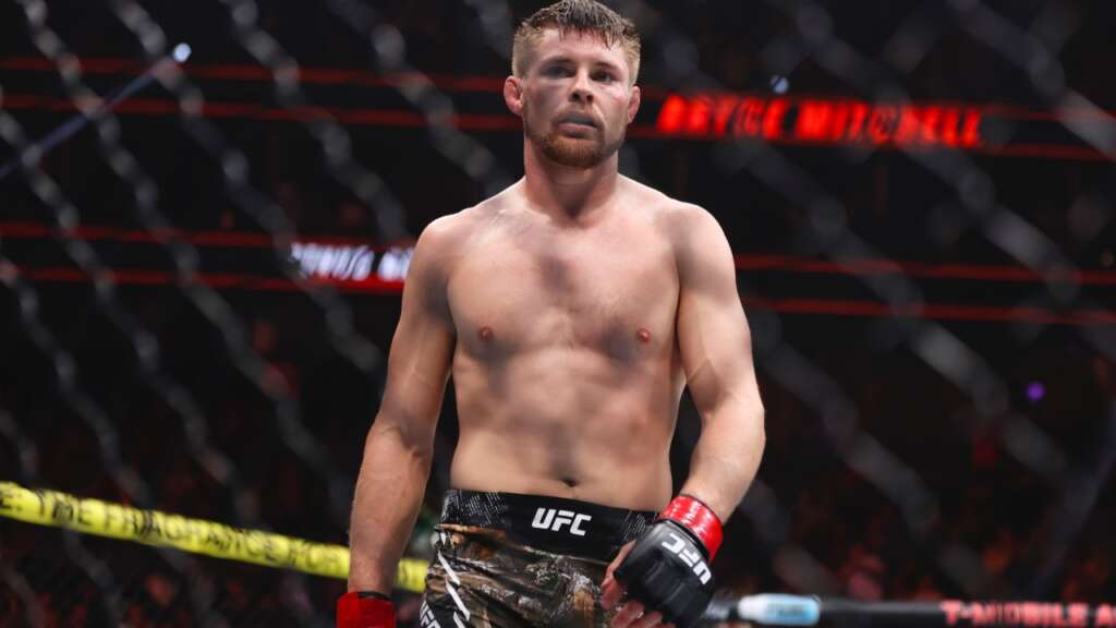 UFC fighter standing in the octagon, shirtless with red gloves and camo shorts, focused expression behind a chain-link fence barrier.