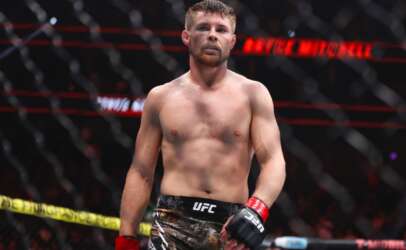 UFC fighter standing in the octagon, shirtless with red gloves and camo shorts, focused expression behind a chain-link fence barrier.