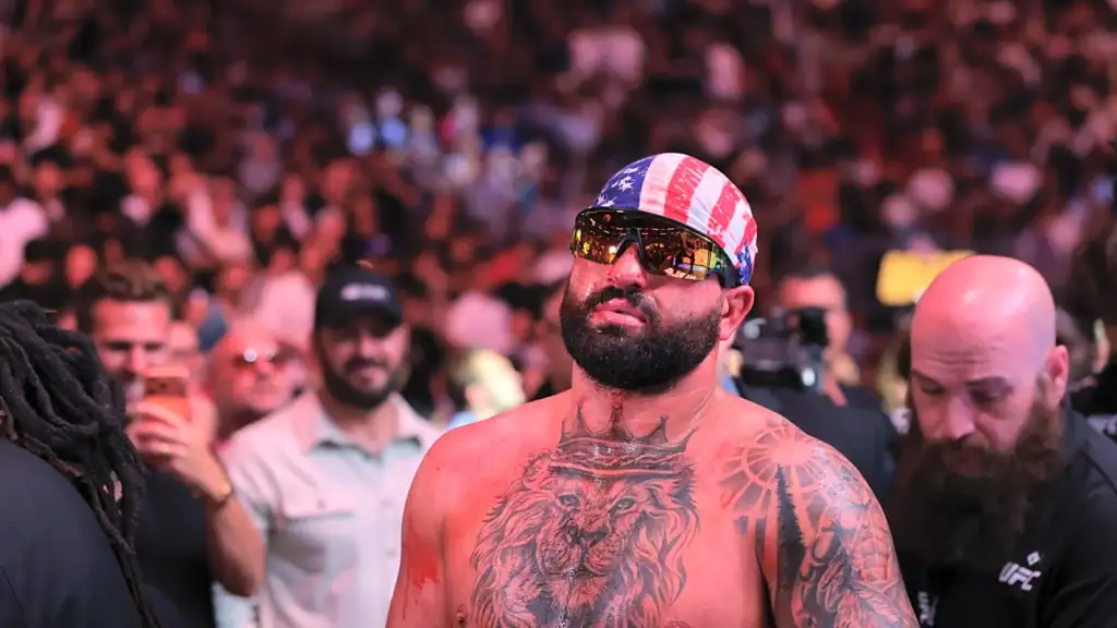 Shirtless, tattooed man with an American flag bandana and mirrored sunglasses standing in a cheering crowd.