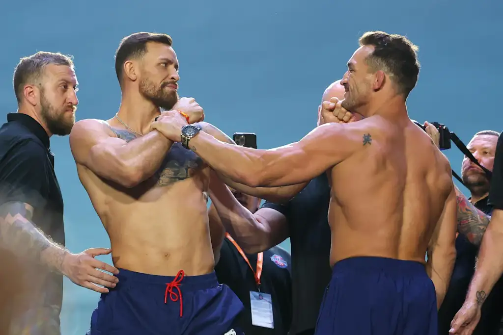Two muscular men in blue shorts grip each other’s wrists in a tense staredown, surrounded by onlookers.