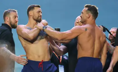 Two muscular men in blue shorts grip each other’s wrists in a tense staredown, surrounded by onlookers.