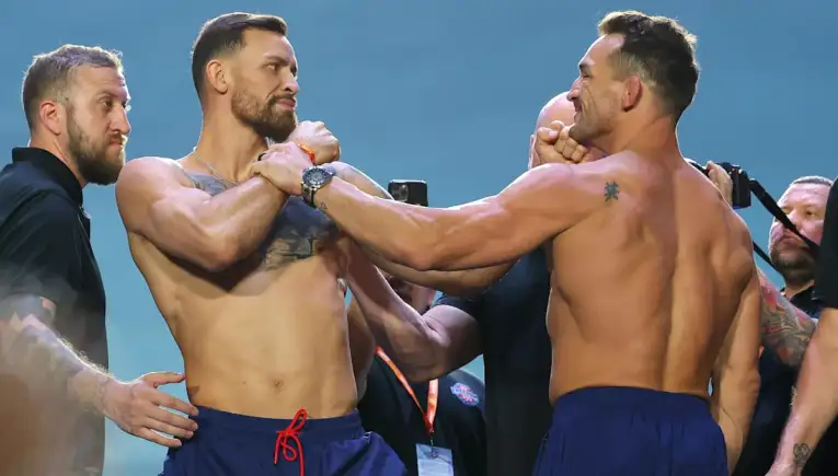 Two muscular men in blue shorts grip each other’s wrists in a tense staredown, surrounded by onlookers.