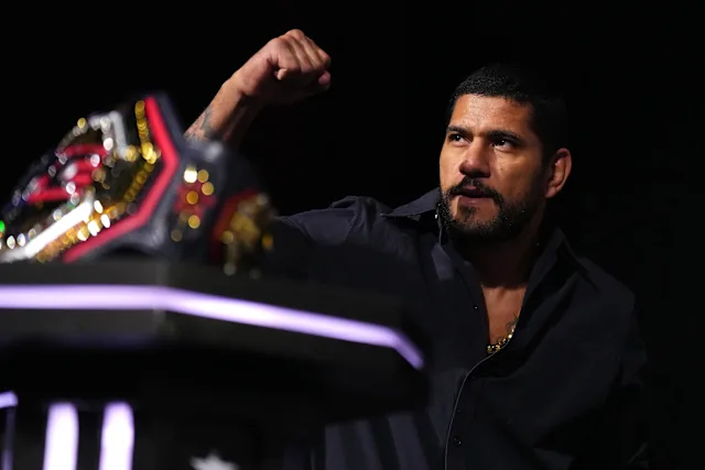 Man in a dark shirt raises a clenched fist in celebration beside a championship belt on a podium or table.