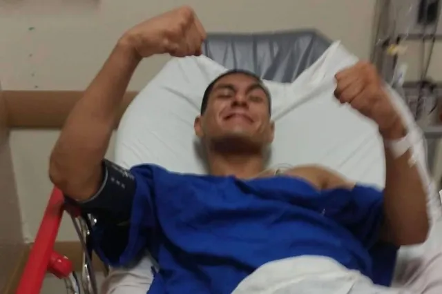Smiling man in a blue hospital gown lying in a bed, giving two thumbs up and showing a bandaged arm in a medical setting.